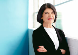 Professor Faye Mishna stands beside a window, smiling with arms crossed