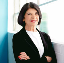Professor Faye Mishna stands beside a window, smiling with arms crossed