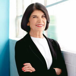 Professor Faye Mishna stands beside a window, smiling with arms crossed