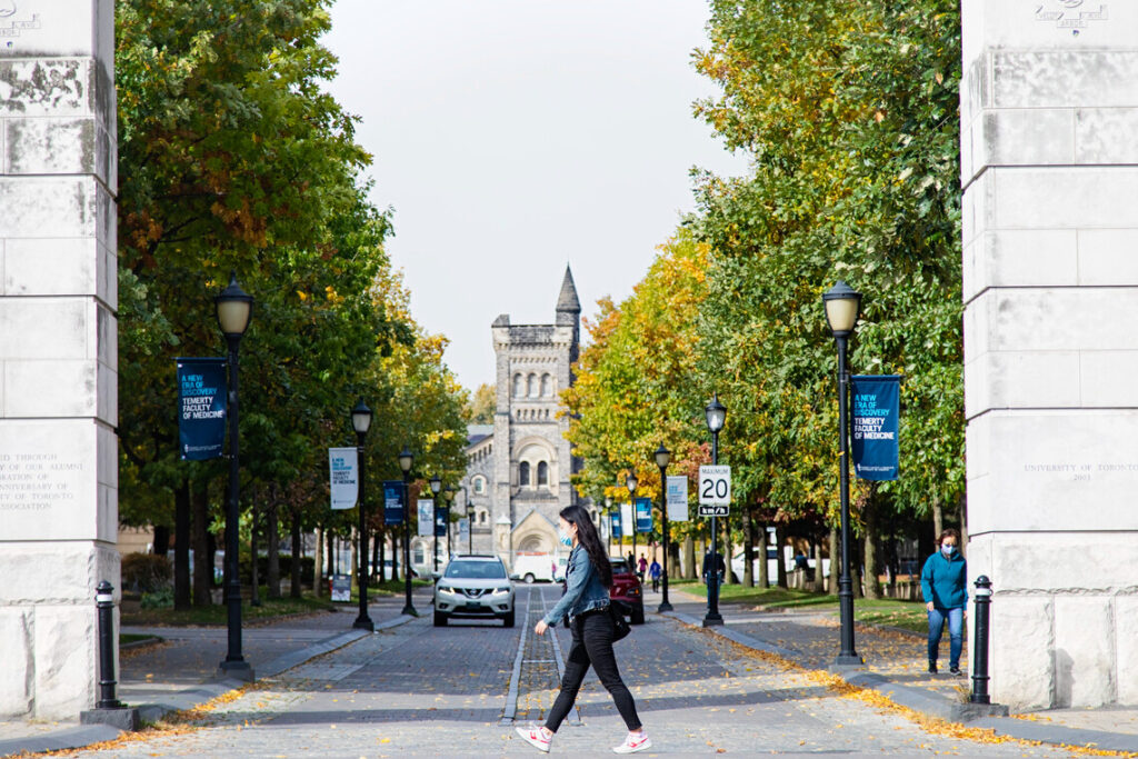 South entrance to the St. George campus with University College in the background and a woman crossing the road in the foreground;.