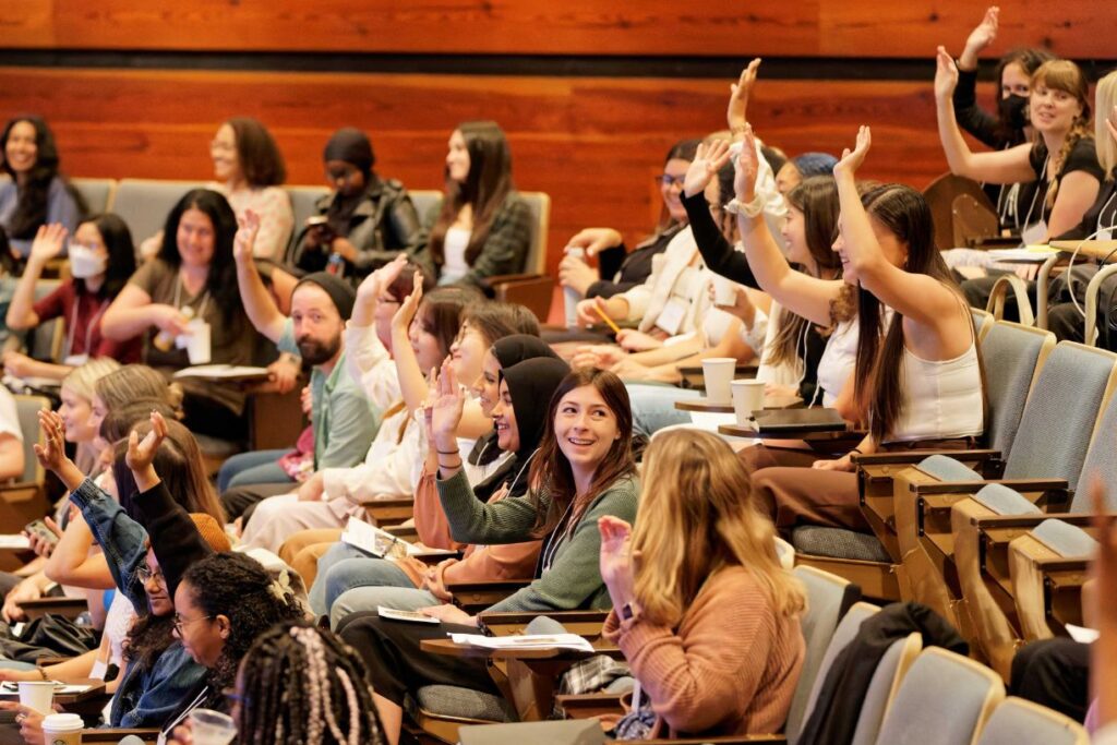 Smiling students in a lecture hall during orientation. Half the student have their hands raised. 