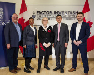 Group photo of Professor David Brennan, Dean Charmaine Williams, the Honourable Carolyn Bennett, Minister of Mental Health and Addictions and Associate Minister of Health, Michael Kwag, Director, CBRC, and Jack Lawrence, participant in the Investigaytors program. Posed in front of FIFSW's logo, flanked by Canadian flags. U of T banner to the left.