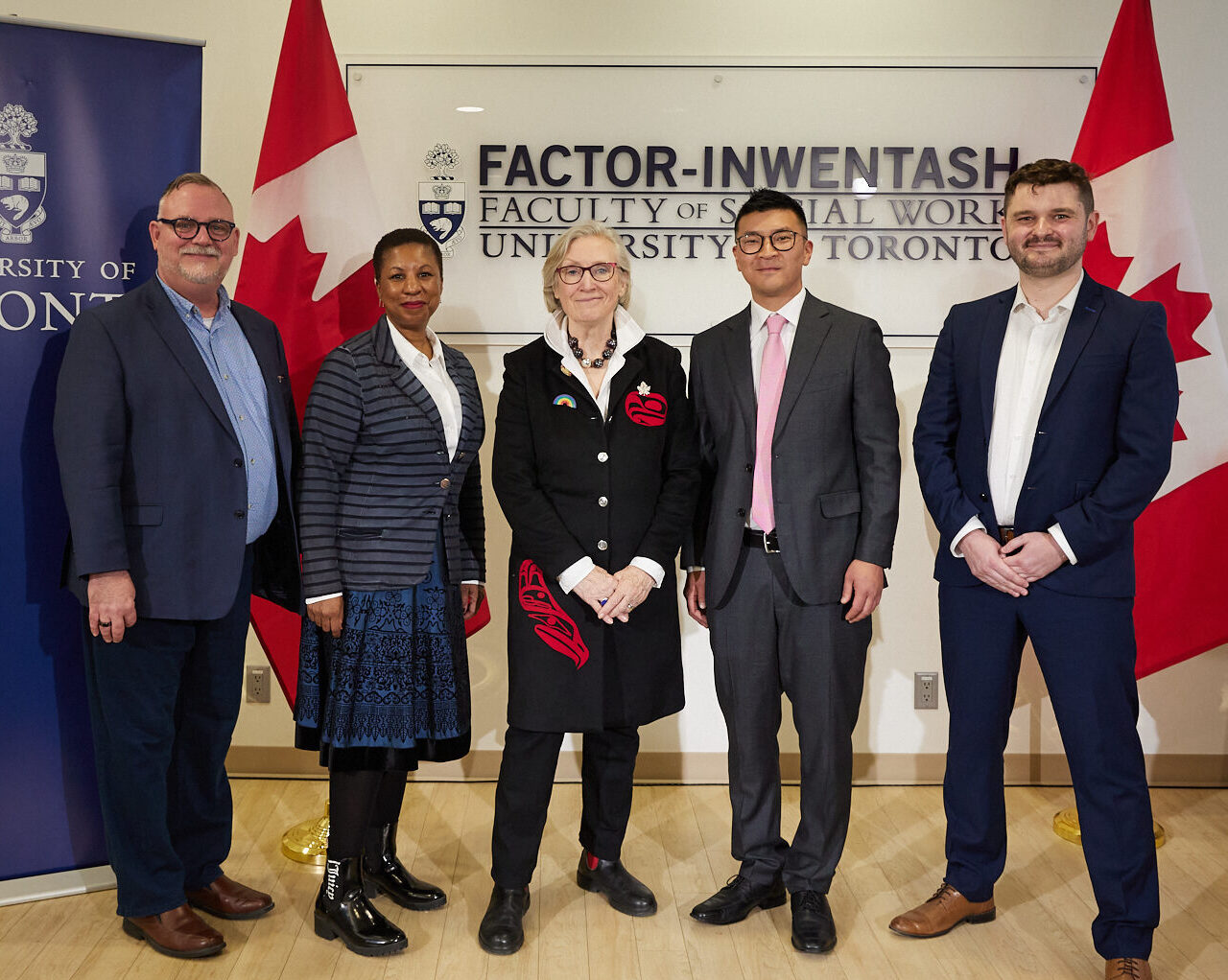 Group photo of Professor David Brennan, Dean Charmaine Williams, the Honourable Carolyn Bennett, Minister of Mental Health and Addictions and Associate Minister of Health, Michael Kwag, Director, CBRC, and Jack Lawrence, participant in the Investigaytors program. Posed in front of FIFSW's logo, flanked by Canadian flags. U of T banner to the left.