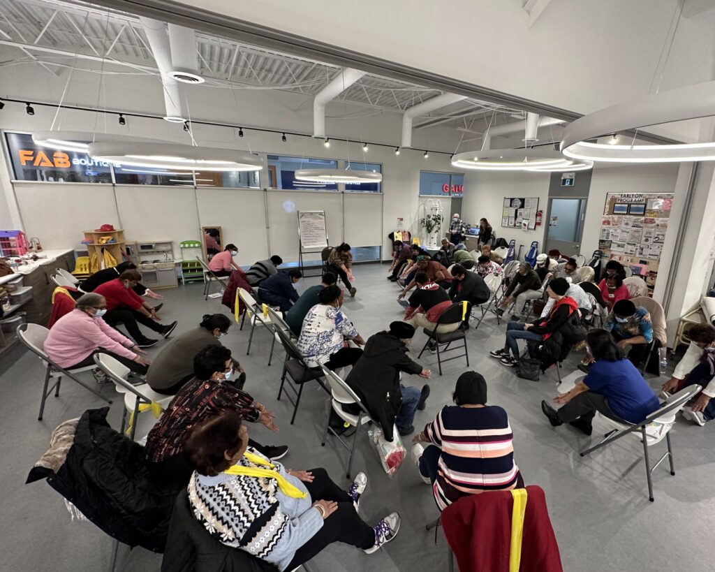 Group of 20+ seniors at the Jane/Finch centre reach for their toes while sitting on chairs. The activity is led by a younger student at the front of the large activity room.
