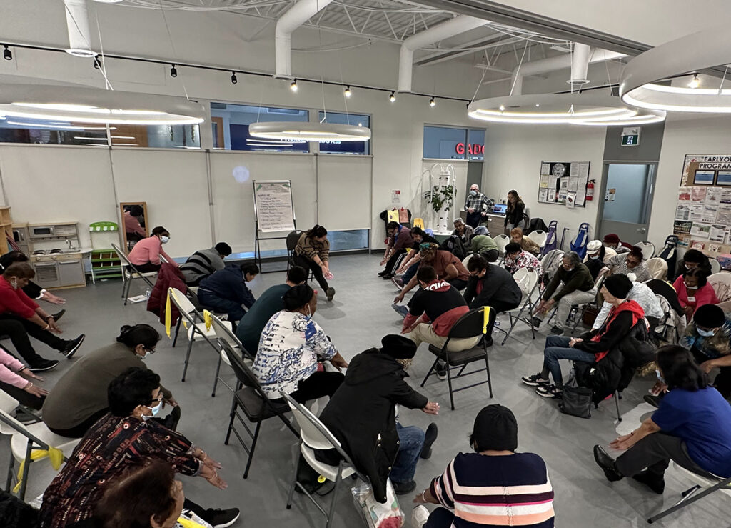 Group of 20+ seniors at the Jane/Finch centre reach for their toes while sitting on chairs. The activity is led by a younger student at the front of the large activity room.