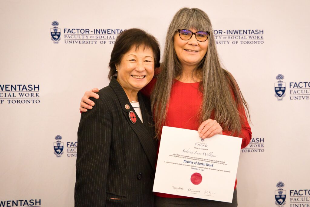 Sabrina Williams poses for a photo with her arm around Judy San. Sabrina is holding her MSW degree. They are standing infant of FIFSW's step-and-repeat backdrop which is white with a blue FIFSW logo repeated across it.