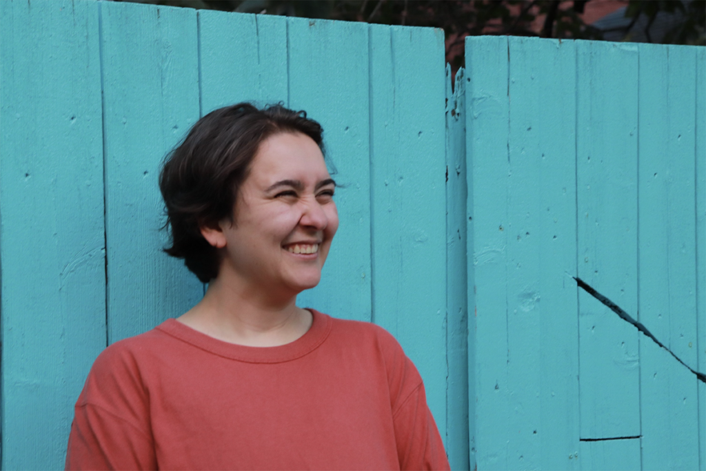 Syd Gilcrest stands against a teal wood fence wearing a red shirt