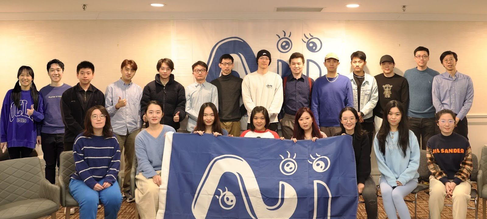 AJI Lab group photo. Members seated in the front row are holding a blue flag with the AIJ Lab logo.