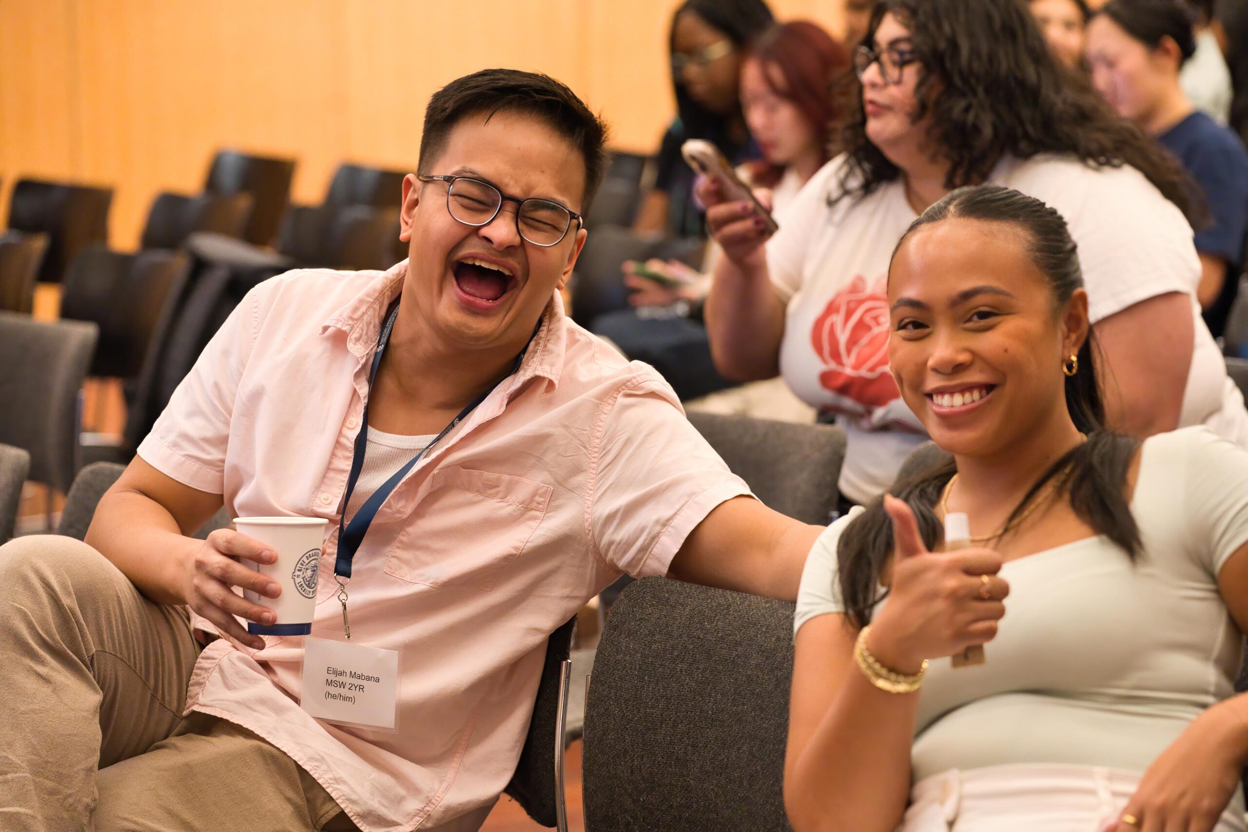 two students seated at a conference pose for a photo. One is laughing, the other is giving a thumbs up.