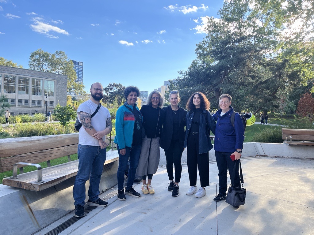 6 people sanding next to a bench with trees and buildings in the background on a sunny day