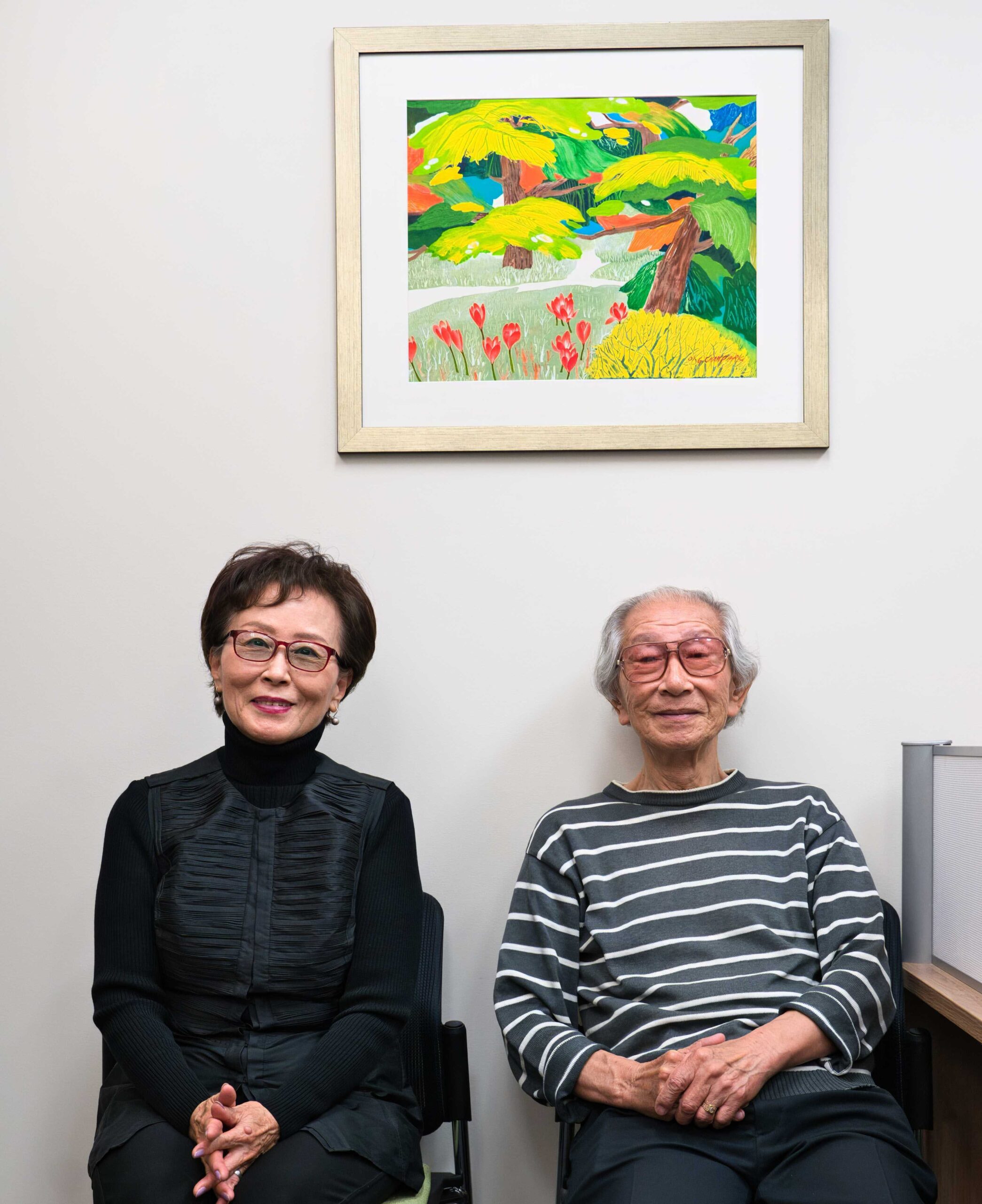 Cha Hee and Suk Kang Park, seated. A colourful painting of Rees and flowers hands on the wall above them.