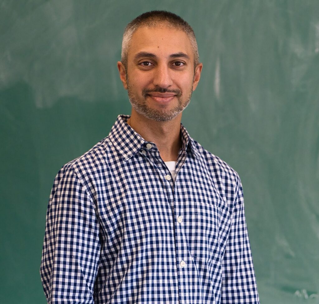 Amar Ghelani, weating a dark blue and white checkered shirt and standing in front of a chalk board