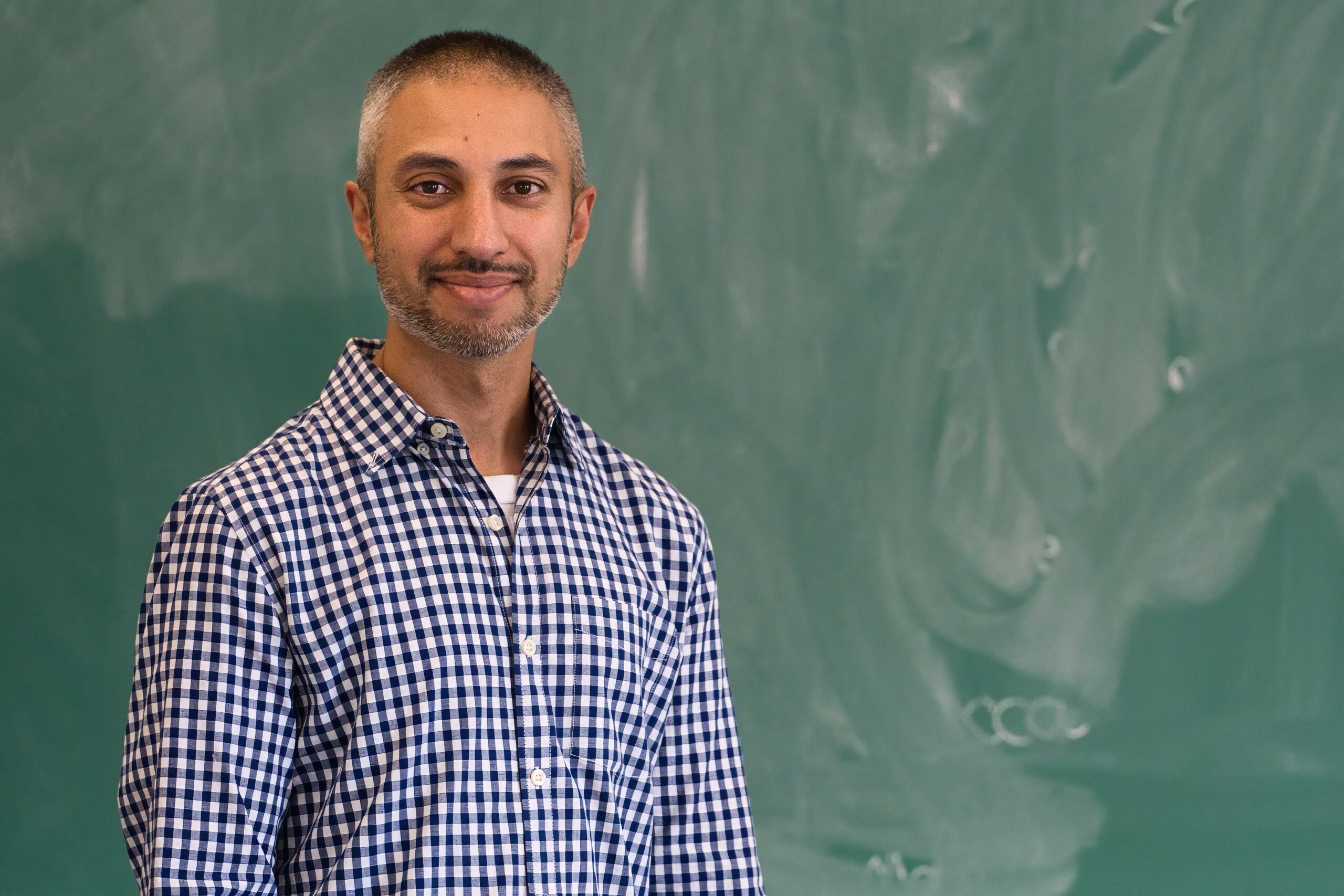 Amar Ghelani, weating a dark blue and white checkered shirt and standing in front of a chalk board