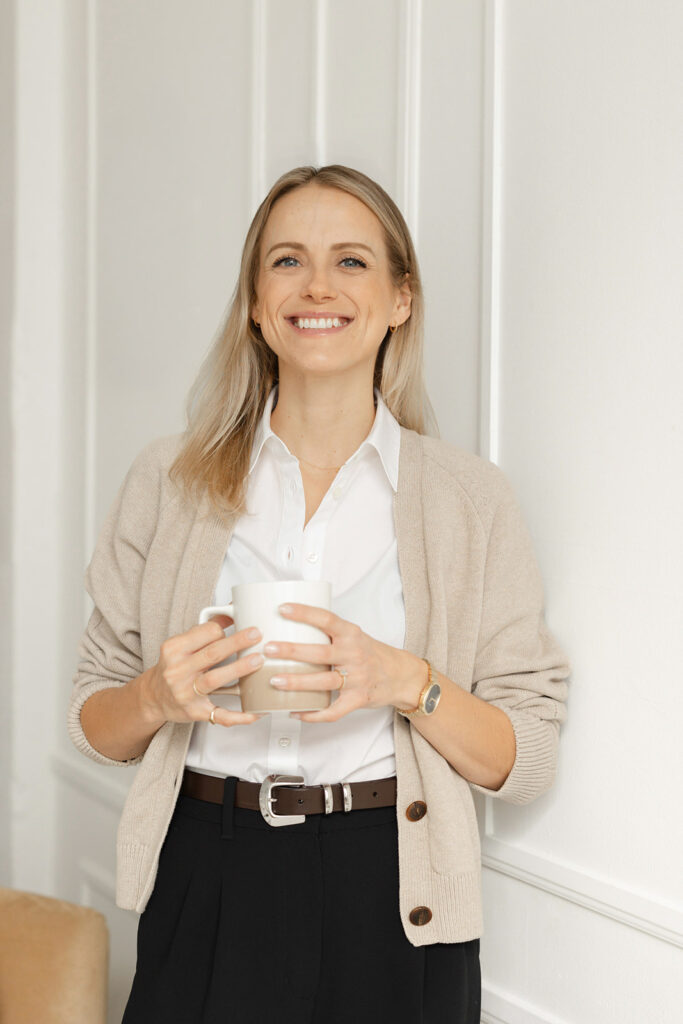 Jenn Shields standing inferno of a white wall wearing a beige cardigan and holding a white coffee cup.