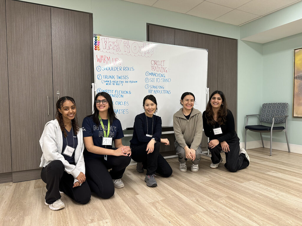 Four women pose for a photo kneeling in front of a white board that lists the exercises that will be done as part of Talk It Out Work It Out 