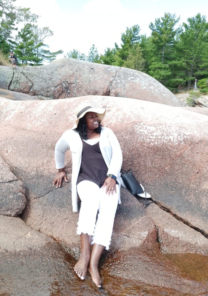 Agnes Nambalirwa wearing a sun hat and leaning against a rock on the shore, her feet in the lake. Trees are in the background.