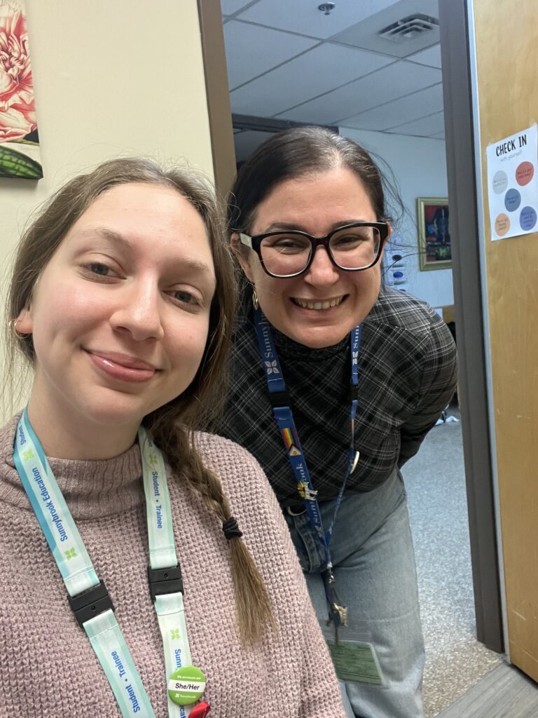 Mayra and Inna wearing lanyards, smile fort a selfie in an office.